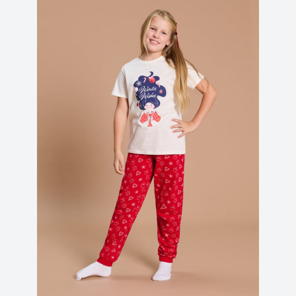 A young girl modeling a two-piece pajama set consisting of a cream-colored t-shirt with a 'Winter Wonderland' graphic and red pants with a white heart pattern, set against a solid beige studio background.