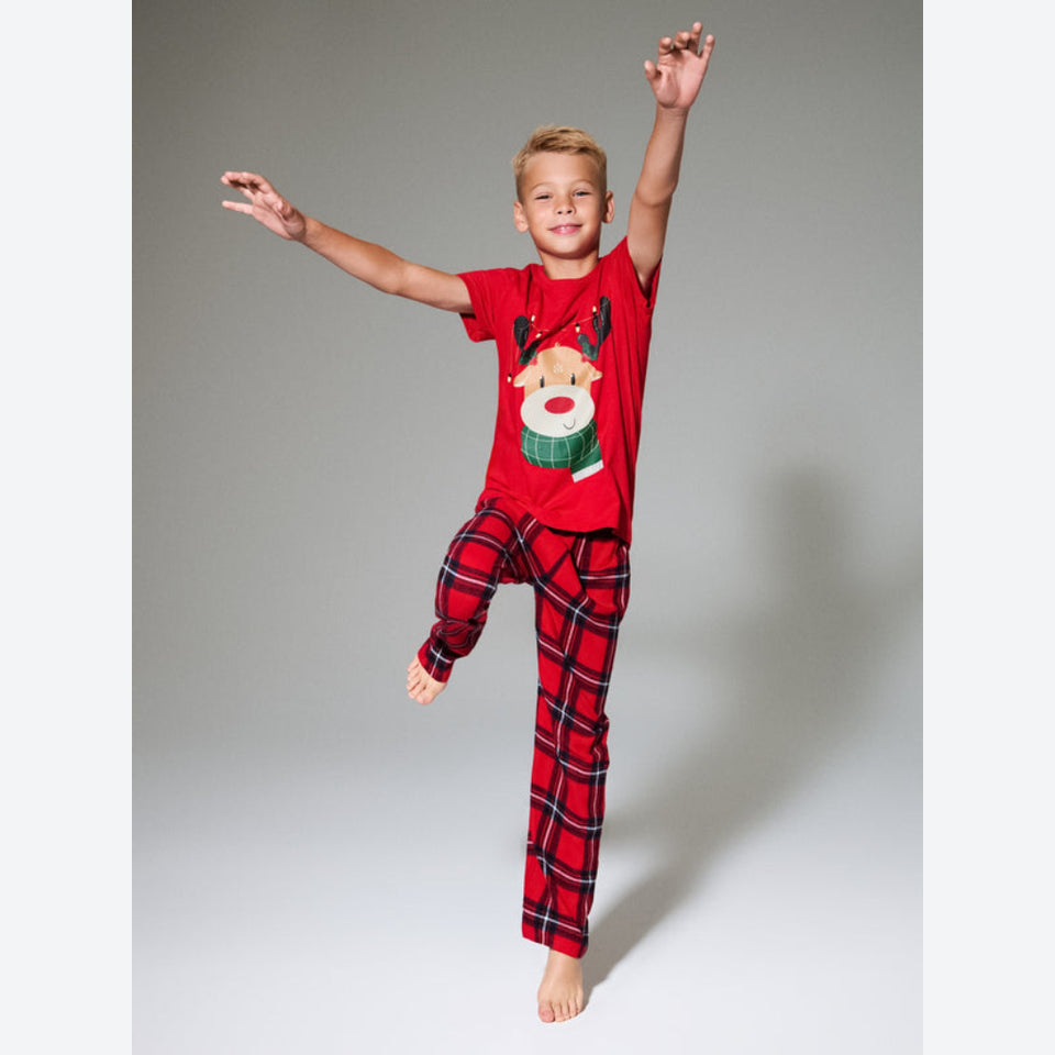 A young boy wearing a red Christmas pajama set with a reindeer graphic on the t-shirt and red-and-black plaid pants, posing dynamically against a neutral gray studio background.
