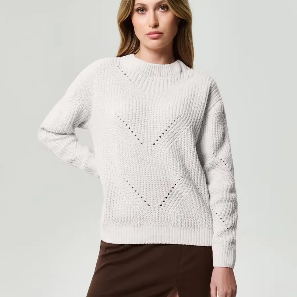 A woman wearing a cream-colored ribbed knit sweater with textured geometric patterns, paired with a brown skirt, against a light gray studio background.