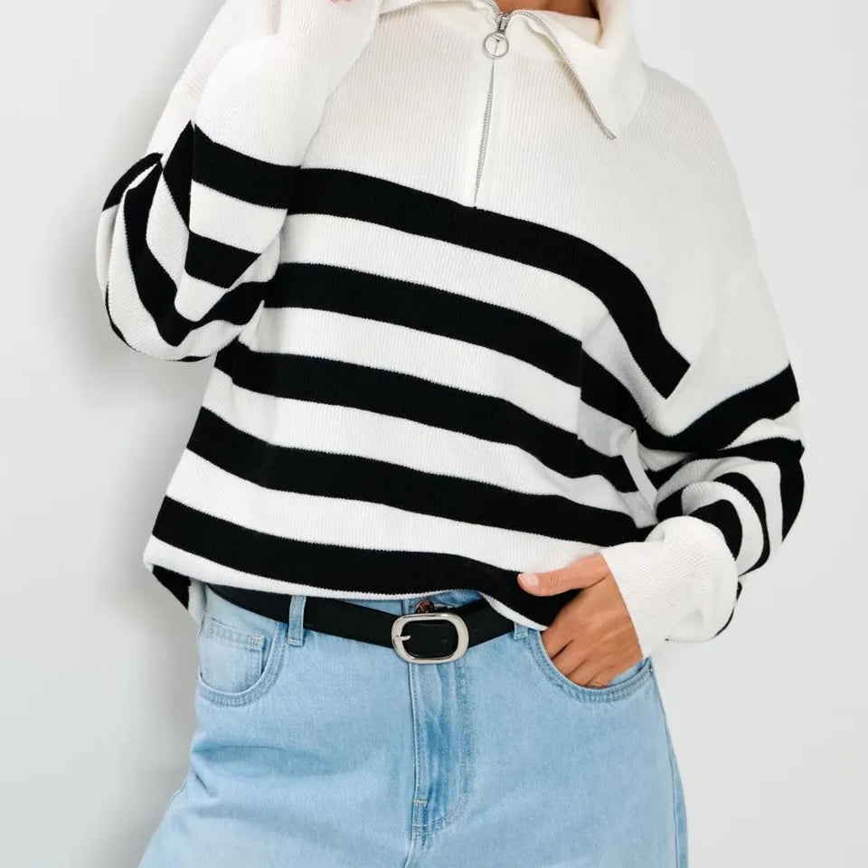 A person wearing a black and white horizontally striped half-zip knit sweater with a white collar, paired with light blue jeans and a black belt.