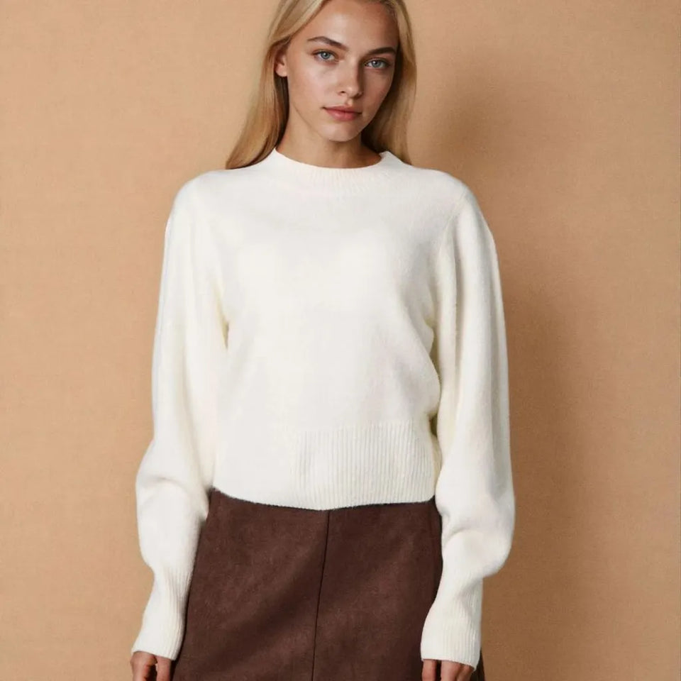 A woman wearing a cream-colored knit sweater and a brown skirt, standing against a solid beige background.