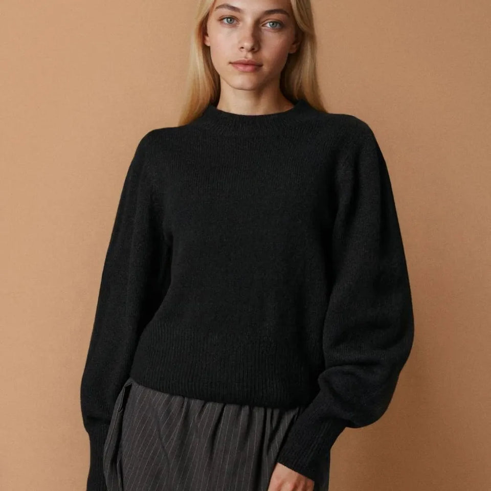 A woman wearing a black knit crewneck sweater with voluminous balloon sleeves, paired with a dark pleated skirt, set against a solid warm tan background.