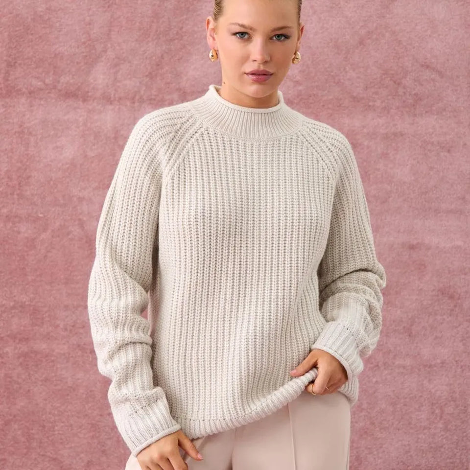 A woman wearing a chunky knit cream-colored mock neck sweater, standing against a textured dusty pink background.