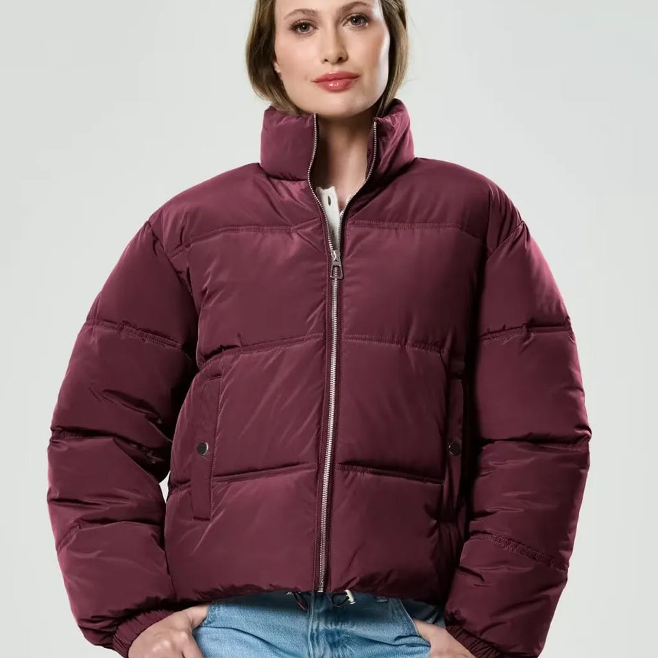 A woman wearing a cropped, quilted puffer jacket in a deep burgundy or maroon color, paired with blue jeans, against a light gray studio background.