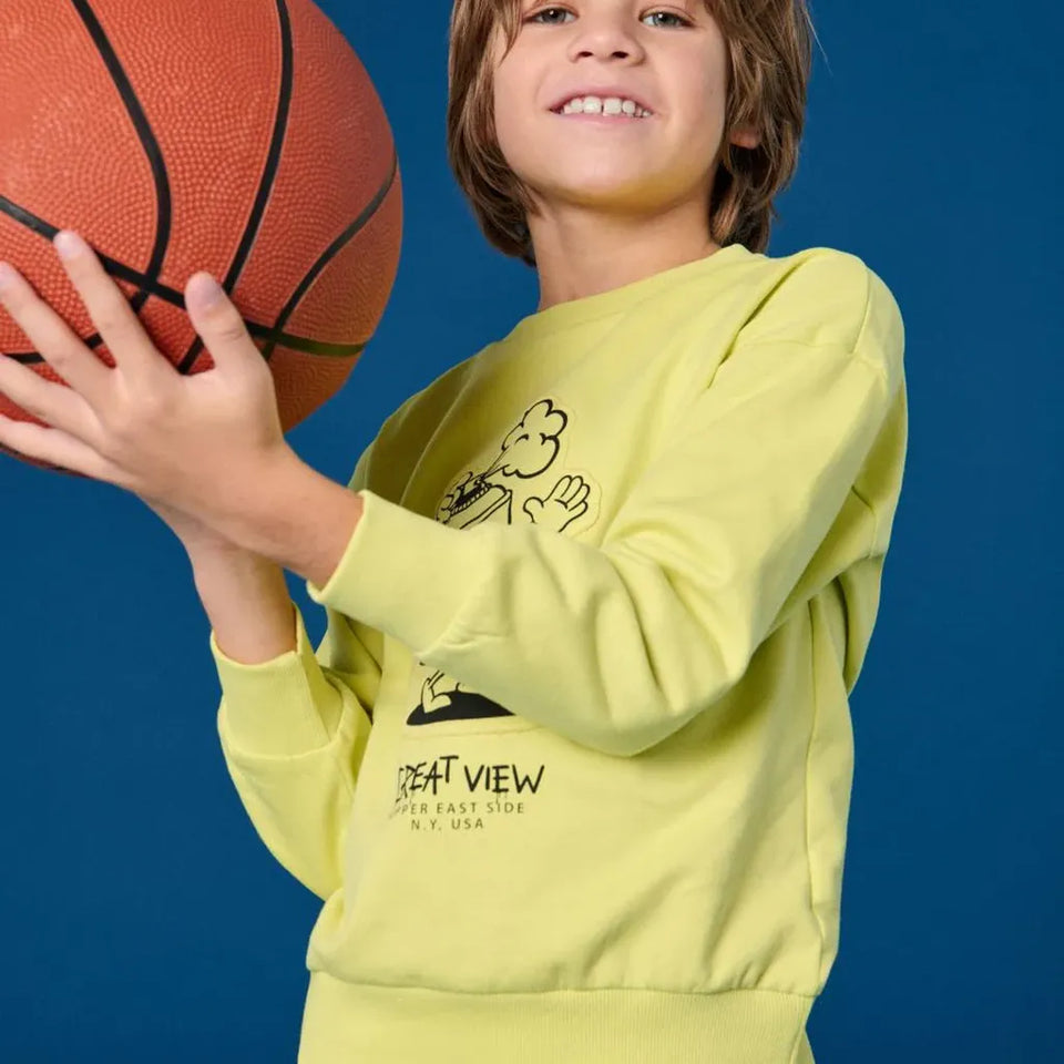 A smiling boy holding a basketball, wearing a light yellow-green long-sleeve sweatshirt with a black cartoon graphic and 'SOUTH VIEW' text, posed against a solid blue background.