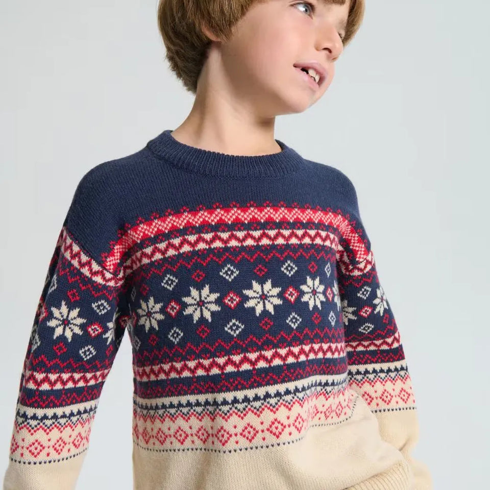 A child wearing a navy blue knit sweater with a red and cream Nordic/Fair Isle pattern across the chest and sleeves, photographed against a light gray studio background.