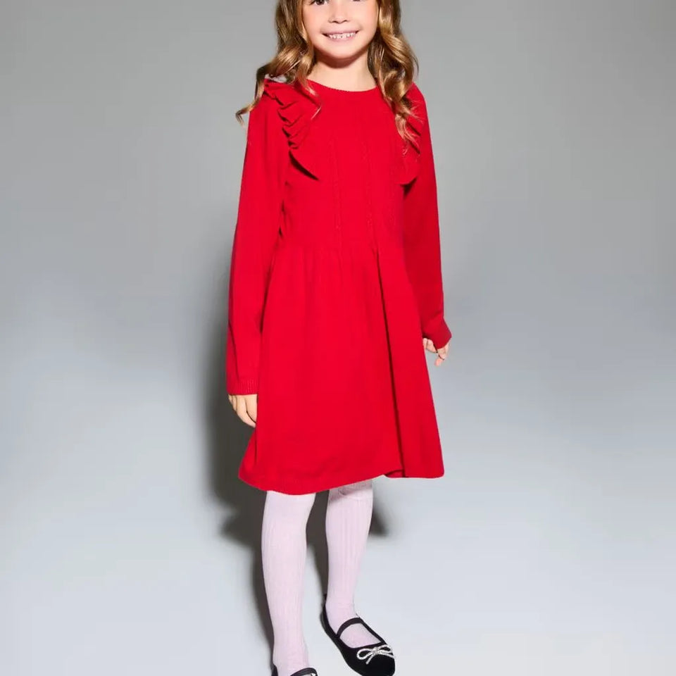 A young girl modeling a bright red long-sleeve dress with ruffle details on the shoulders, paired with light pink tights and black ballet flats, against a neutral gray studio background.