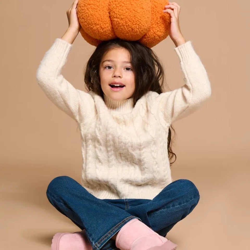 A young girl with long dark hair sits cross-legged on a beige studio background, wearing a cream cable-knit turtleneck sweater, blue jeans, and pink socks. She holds an orange fuzzy pumpkin-shaped pillow above her head with both hands.