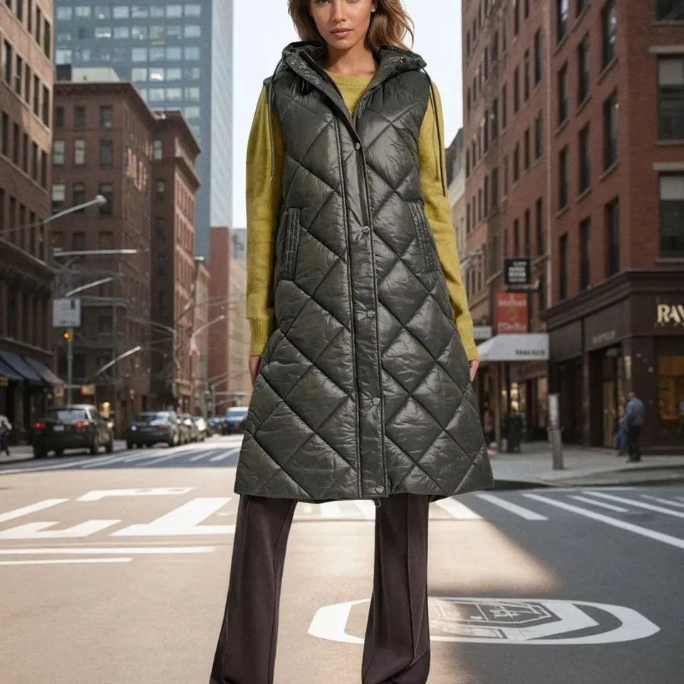 A woman stands on a city street wearing a long diamond-quilted sleeveless puffer vest over a mustard yellow top and dark wide-leg pants.