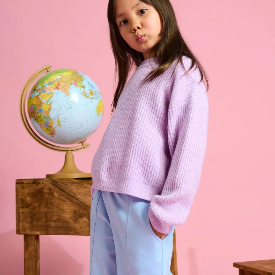 A young girl with long brown hair wearing a lavender waffle-knit sweater and light blue pants, posing next to a globe on a wooden stand against a solid pink background.