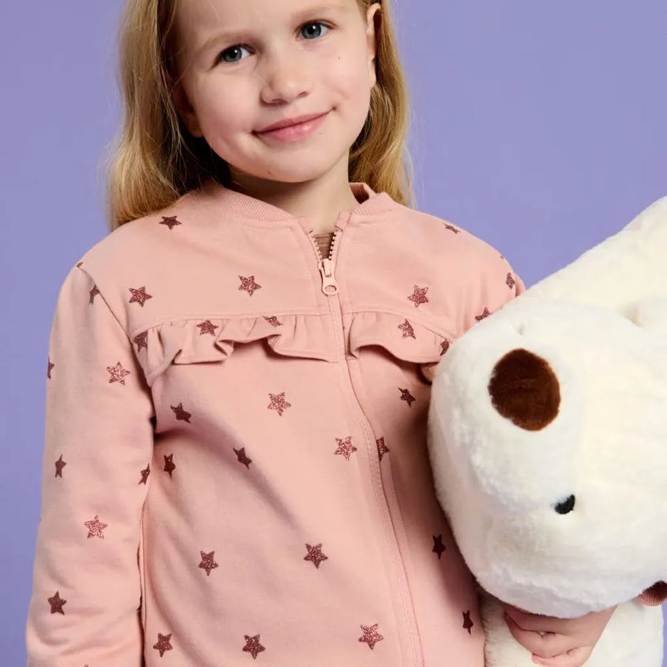 A young girl with blonde hair smiling while wearing a pink zip-up jacket with embroidered brown stars and holding a white plush toy against a solid lavender background.