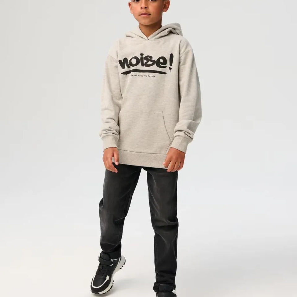 A child wearing a light heather gray hoodie with a black 'noise!' graphic on the chest, paired with dark gray pants and black sneakers, standing against a light gray studio background.