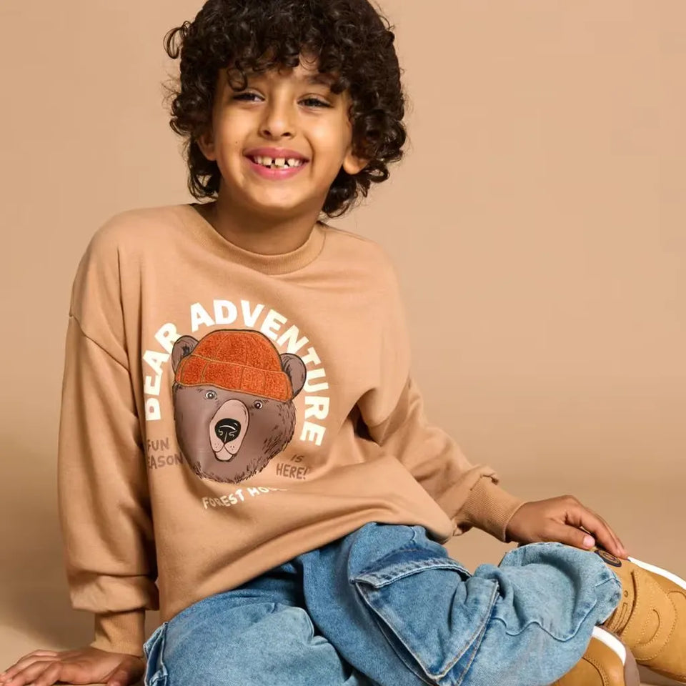 A smiling young boy with curly dark hair sits against a warm beige studio background, wearing a tan crewneck sweatshirt with a bear graphic and the words 'PEAR ADVENTURE' across the front, blue jeans, and tan boots.