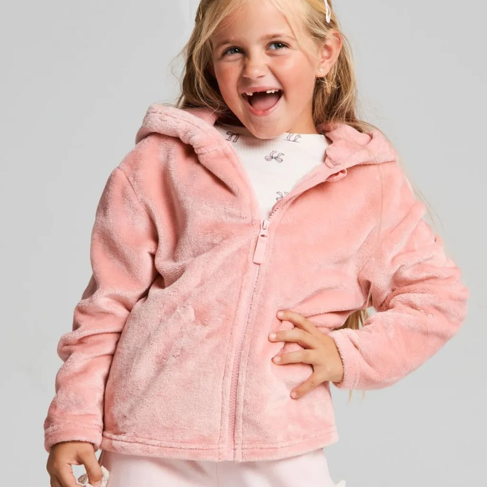 A young girl with blonde hair smiling and laughing while wearing a pink hooded zip-up fleece jacket over a white patterned shirt, set against a plain light gray studio background.