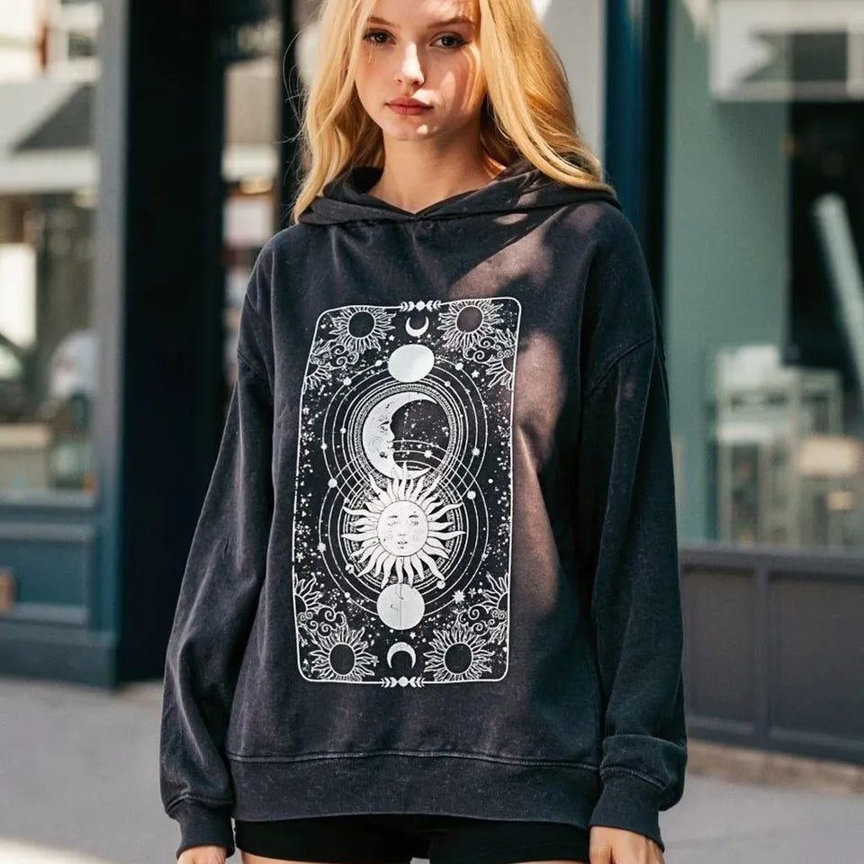 A woman wearing a faded black pullover hoodie with a large white celestial graphic on the front, standing outdoors in front of a blurred storefront.