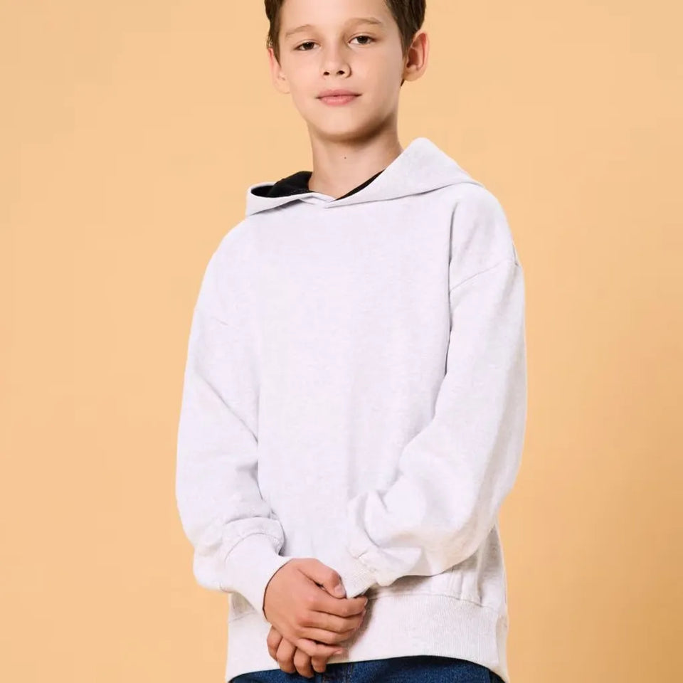 A young boy wearing a light gray oversized pullover hoodie with a black-lined hood, standing against a solid warm beige background.