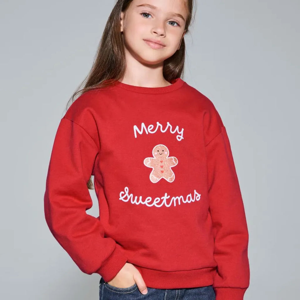 A young girl with long brown hair wearing a red crewneck sweatshirt with 'Merry Sweetmas' and a gingerbread man graphic on the front, paired with blue jeans, against a solid gray studio background.