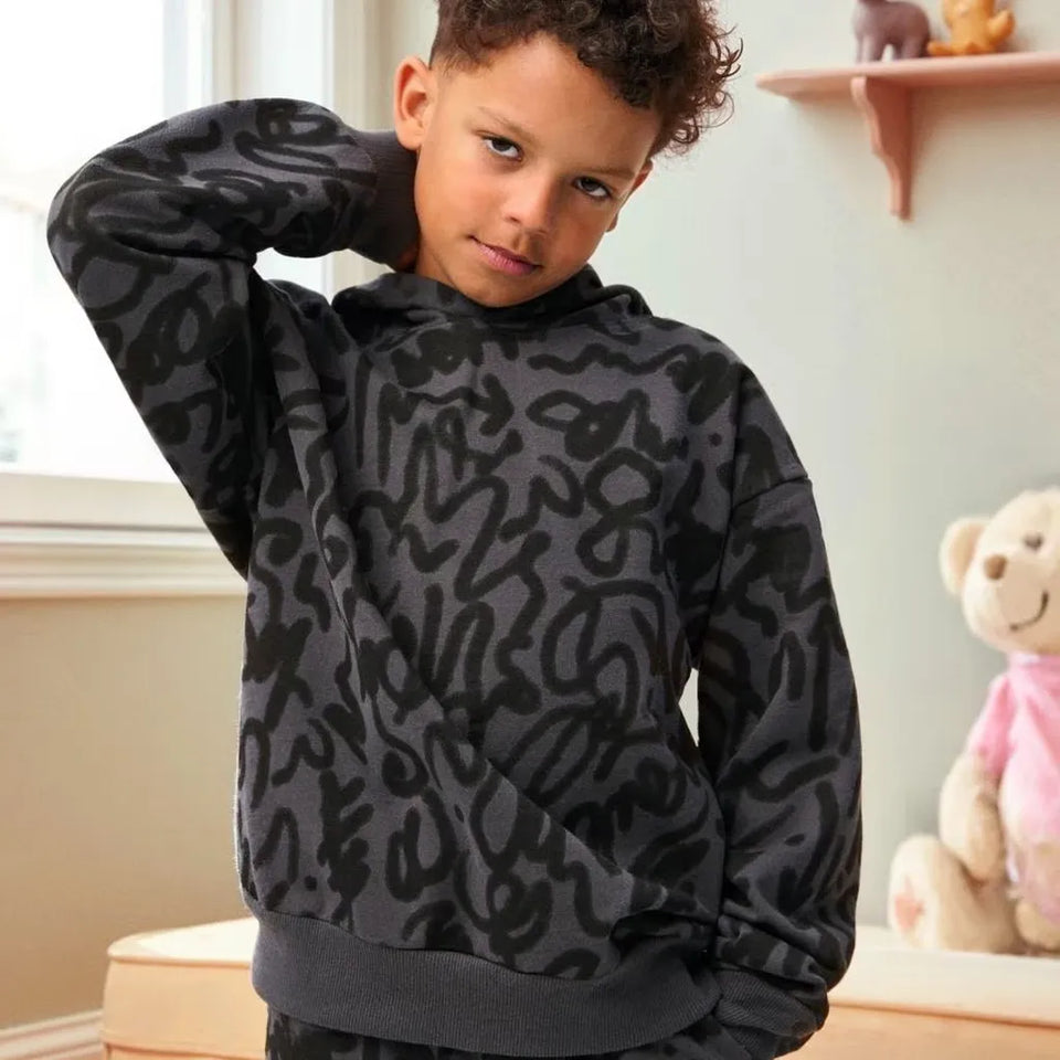 A child wearing a dark gray and black abstract patterned hoodie sweatshirt, standing in a softly lit room with a teddy bear visible in the background.