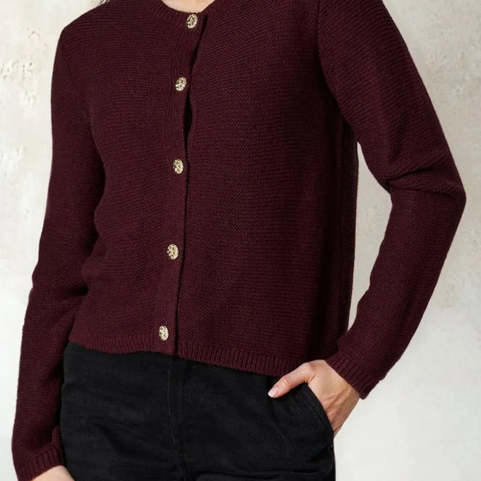 A person wearing a dark burgundy knit cardigan with gold-toned buttons and black pants, standing against a light textured wall.