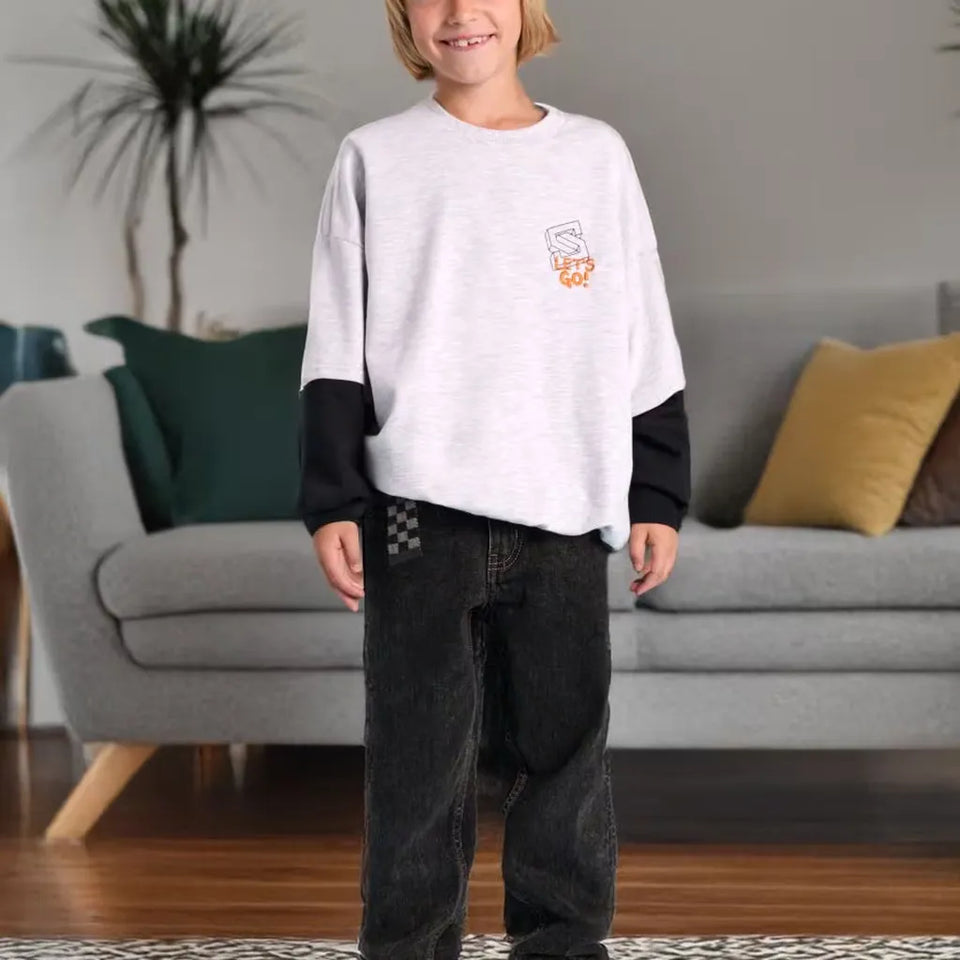 A child wearing a white and black long-sleeve sweatshirt with a small orange and white graphic on the left chest, paired with dark jeans, standing in a living room in front of a gray sofa.