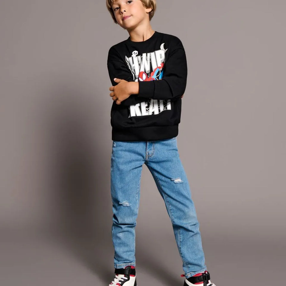 A young boy modeling a black long-sleeve graphic sweatshirt with distressed light blue jeans and black, white, and red sneakers, standing against a solid gray studio background.