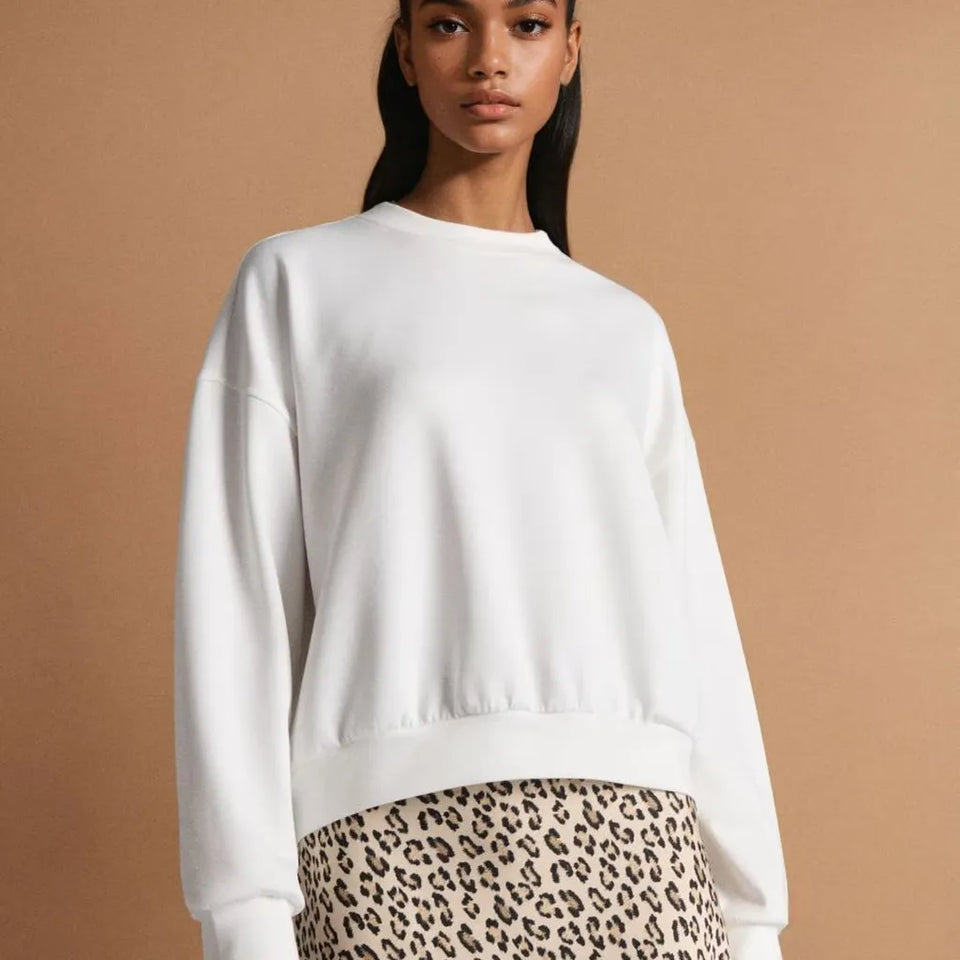A woman wearing a white crewneck sweatshirt and a leopard print skirt, standing against a solid tan background.