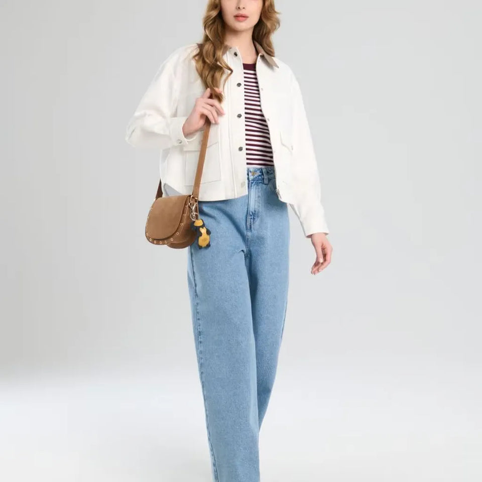 A model wearing a white cropped jacket, a red and white striped t-shirt, light blue wide-leg jeans, and carrying a brown leather shoulder bag with a yellow tassel charm, set against a light gray studio background.