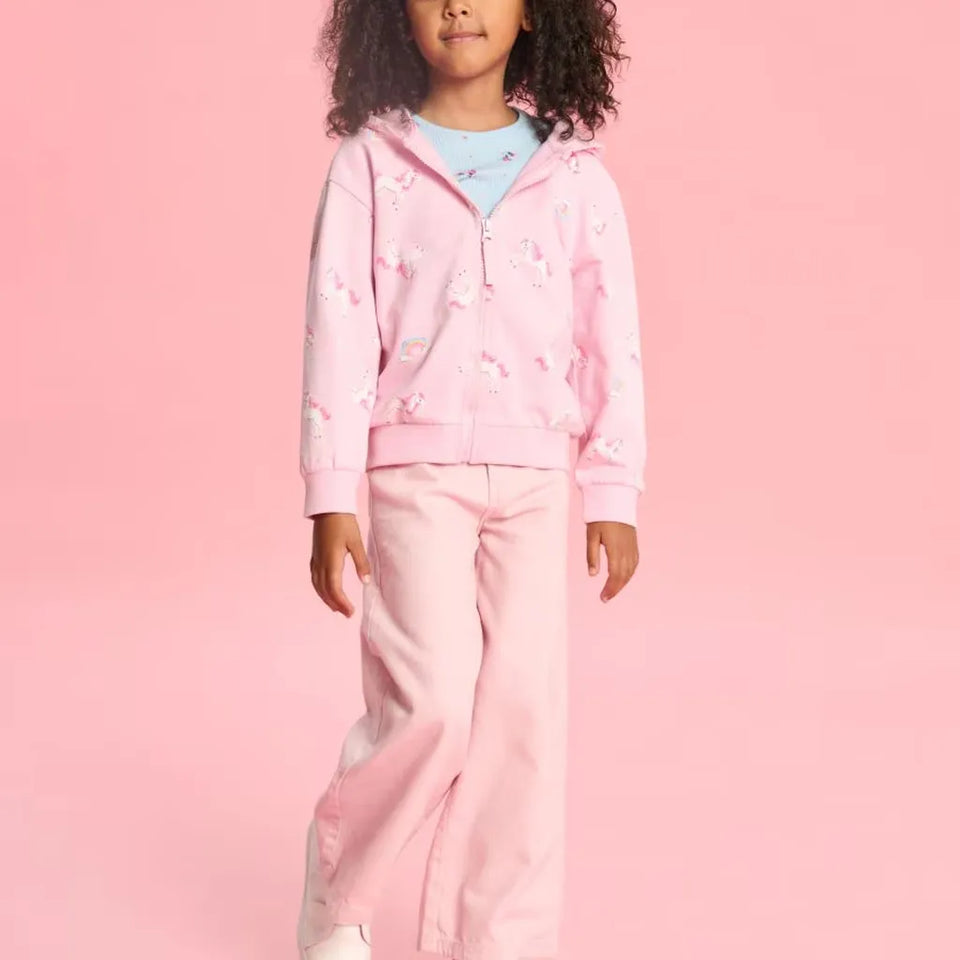 A young girl wearing a pink zip-up hoodie with white unicorn prints and matching pink pants, standing against a solid pink background.