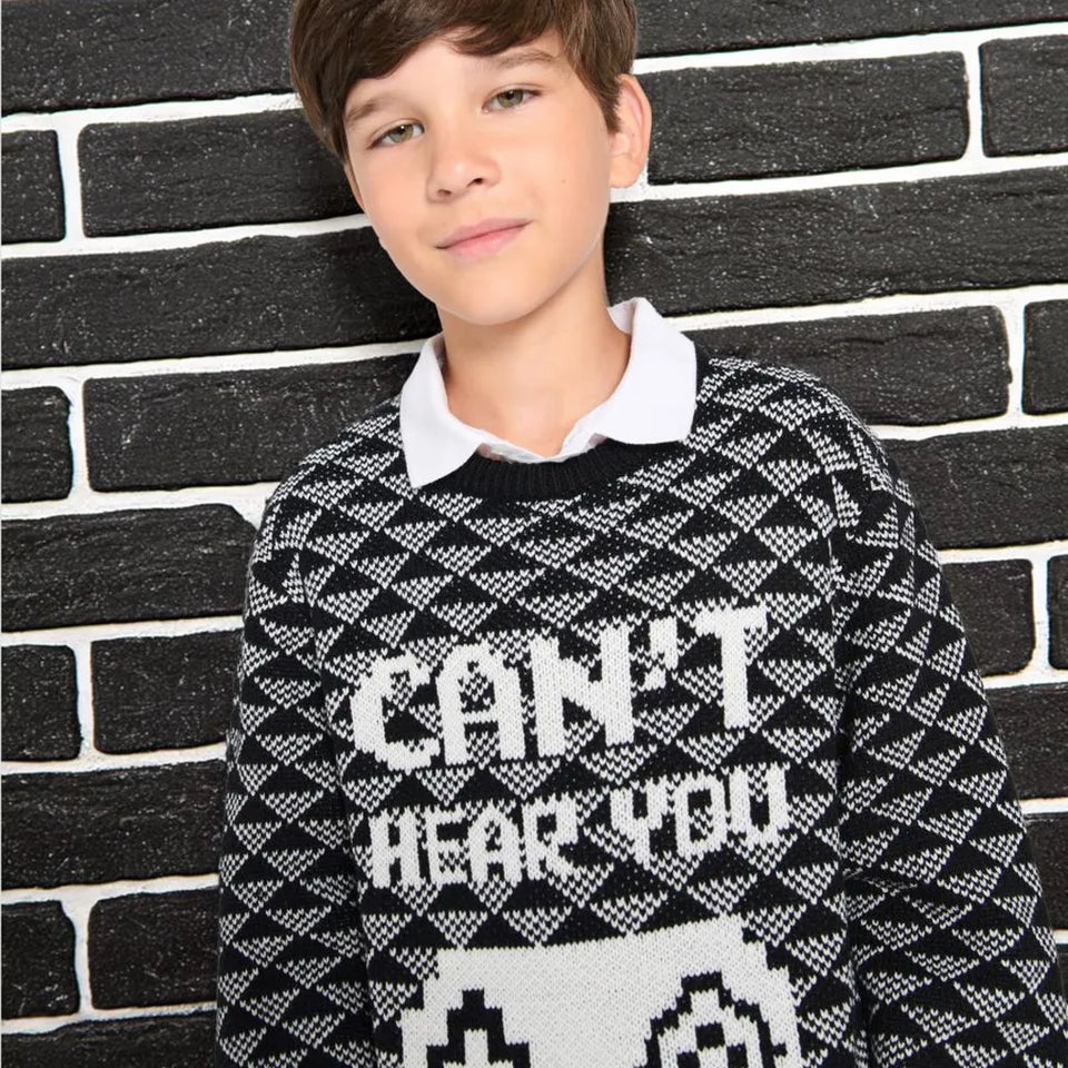 A boy wearing a black and white patterned knit sweater with the text 'CAN'T HEAR YOU' and a pixelated face graphic, layered over a white collared shirt, standing against a black brick wall.