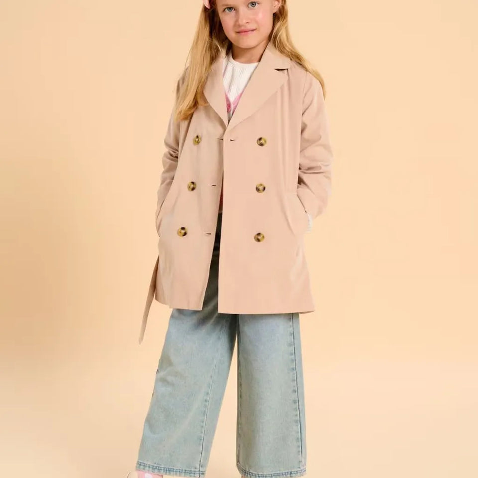 A young girl wearing a light beige double-breasted coat with gold buttons, wide-leg light blue jeans, and a white top, standing against a solid beige background.