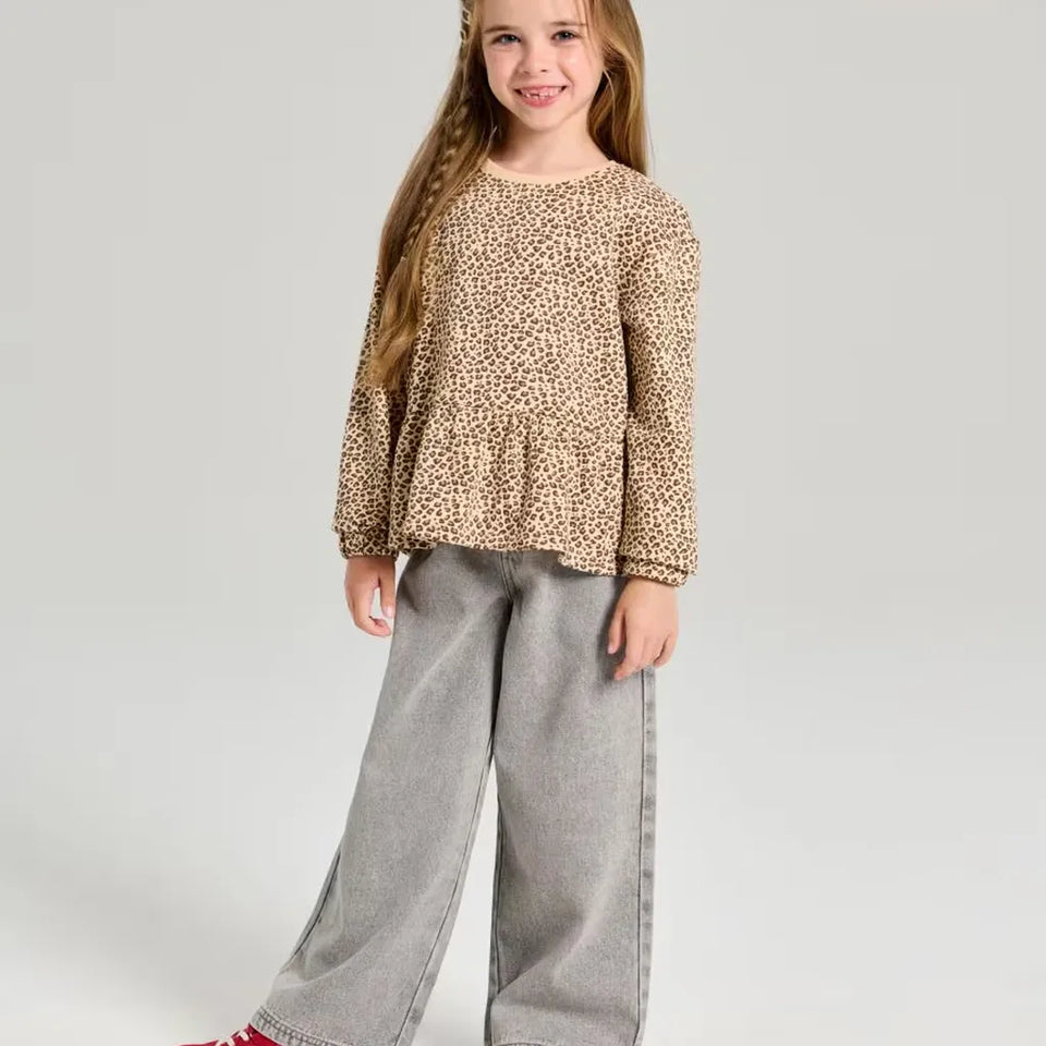 A young girl with long light brown hair stands against a light gray studio background, wearing a beige and brown leopard-print long-sleeve top with a peplum hem and wide-leg gray pants.