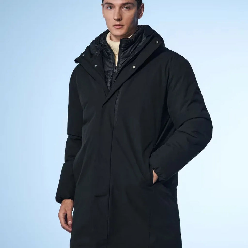 A man wearing a long black hooded puffer parka over a light beige shirt, photographed against a light blue studio background.