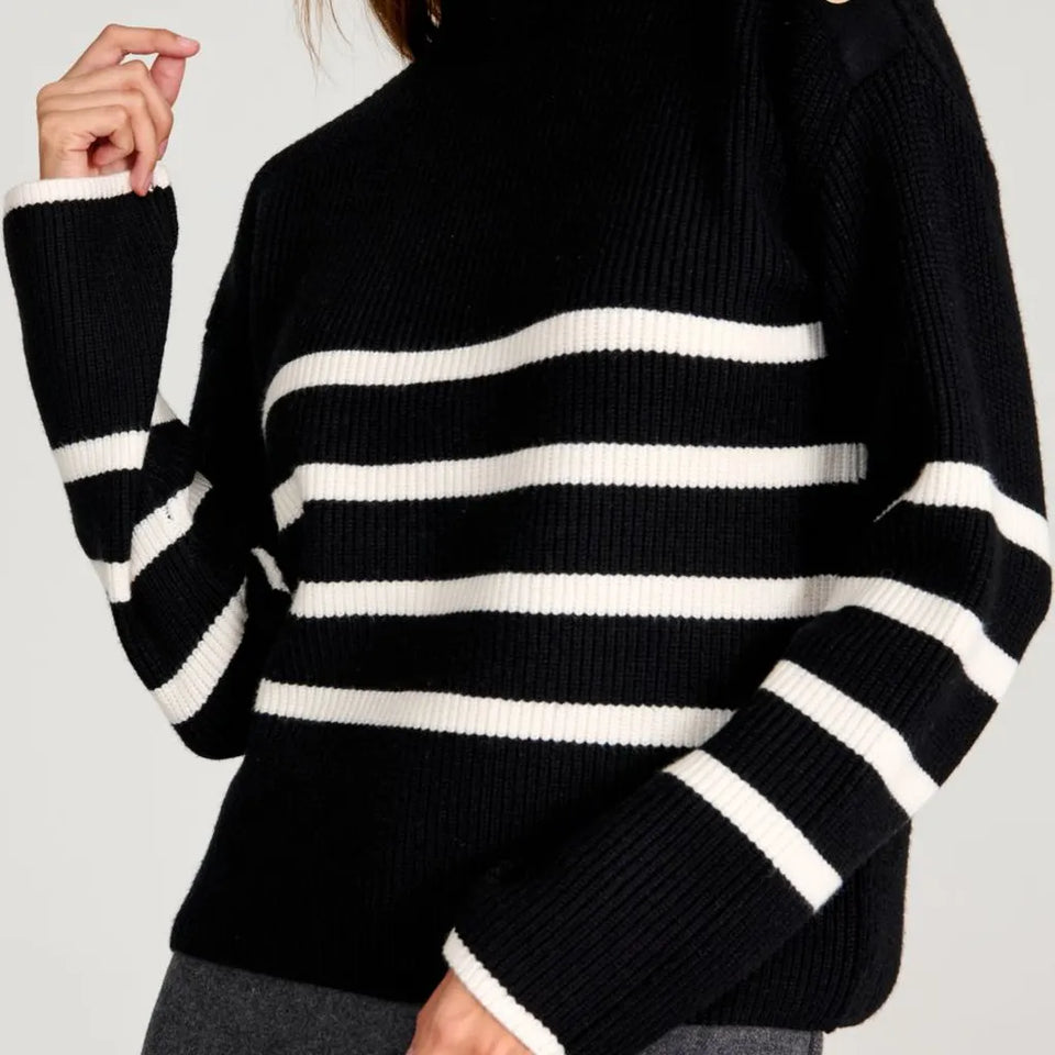 A close-up of a person wearing a black ribbed knit sweater with horizontal white stripes across the chest and sleeves, paired with gray bottoms against a light gray background.