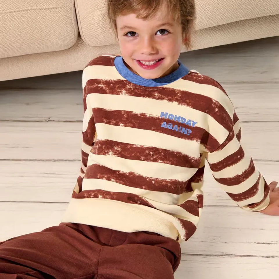 A smiling young boy wearing a brown and cream horizontally striped long-sleeve shirt with a blue collar and 'MONDAY AGAIN?' text on the chest, sitting on a light wood floor in front of a beige sofa.