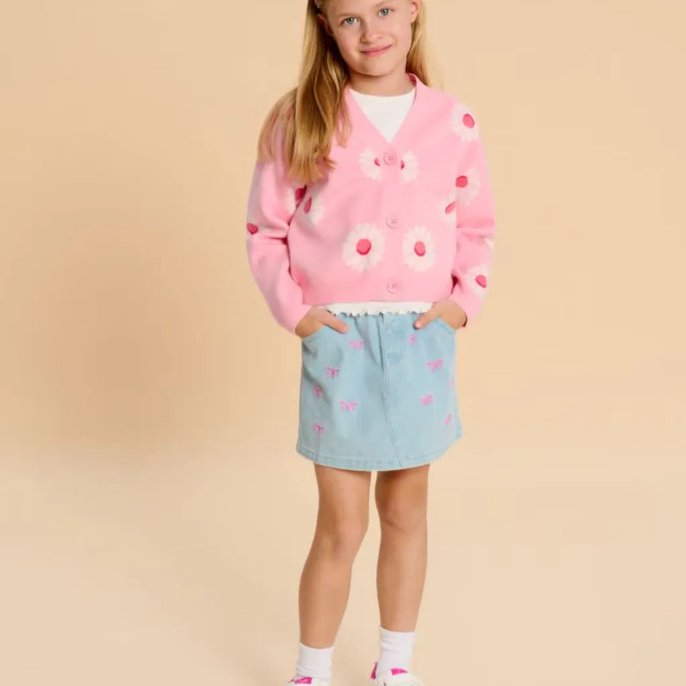 A young girl stands against a solid beige studio background wearing a pink cardigan with white daisy embroidery, a light blue denim skirt with pink bow details, a white t-shirt, white socks, and pink sneakers.