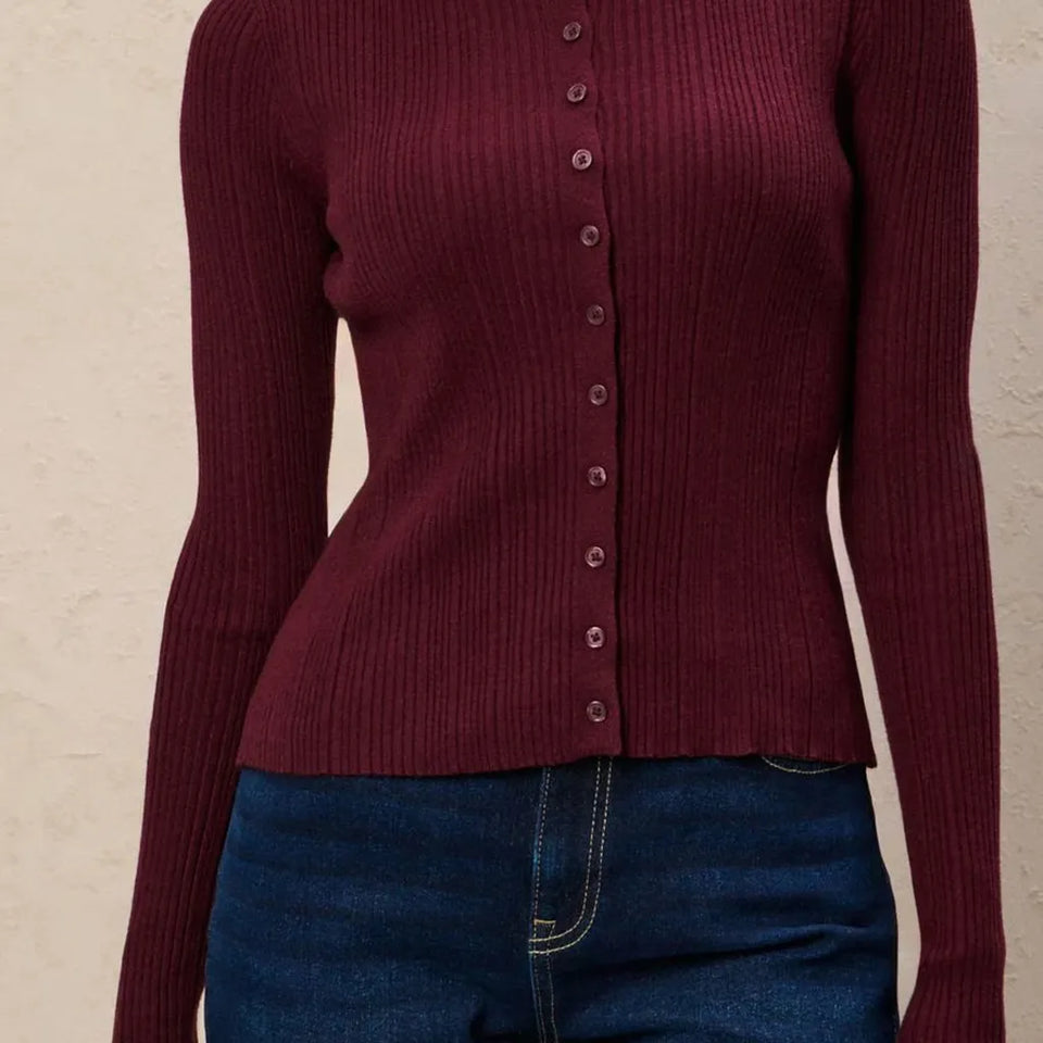 A close-up of a person wearing a ribbed long-sleeve cardigan in deep burgundy with a button-front closure, paired with blue jeans against a light textured wall.