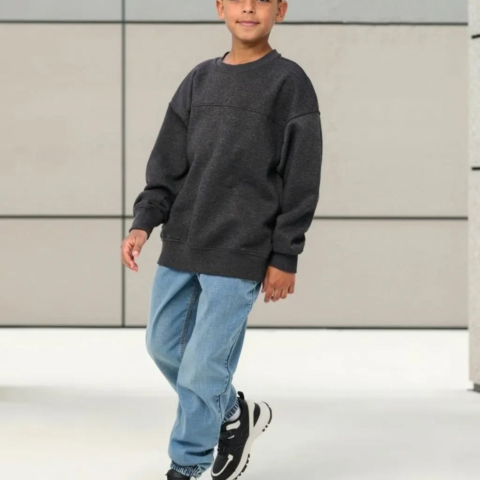 A child wearing an oversized dark charcoal gray crewneck sweatshirt, light blue jeans, and black sneakers, standing against a modern light gray paneled wall.