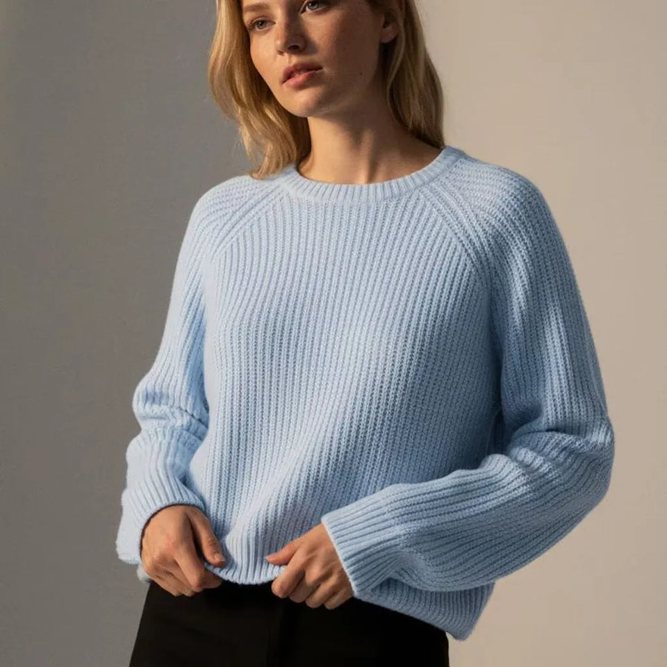 A woman wearing a light blue ribbed knit sweater with long raglan sleeves, paired with black pants, photographed against a neutral beige studio background.