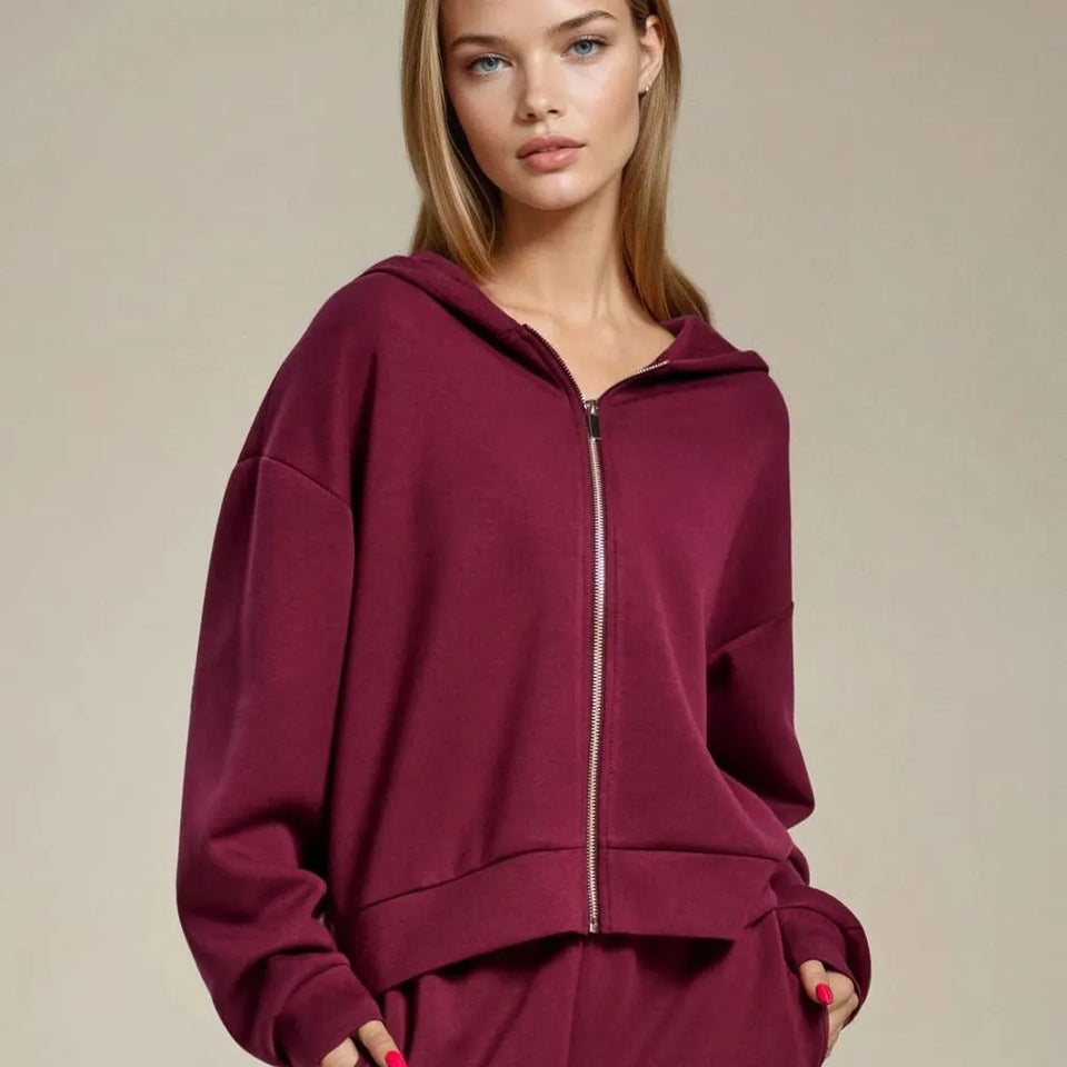 A woman wearing an oversized zip-up hoodie in deep burgundy with a matching pair of sweatpants, photographed against a plain beige studio background.