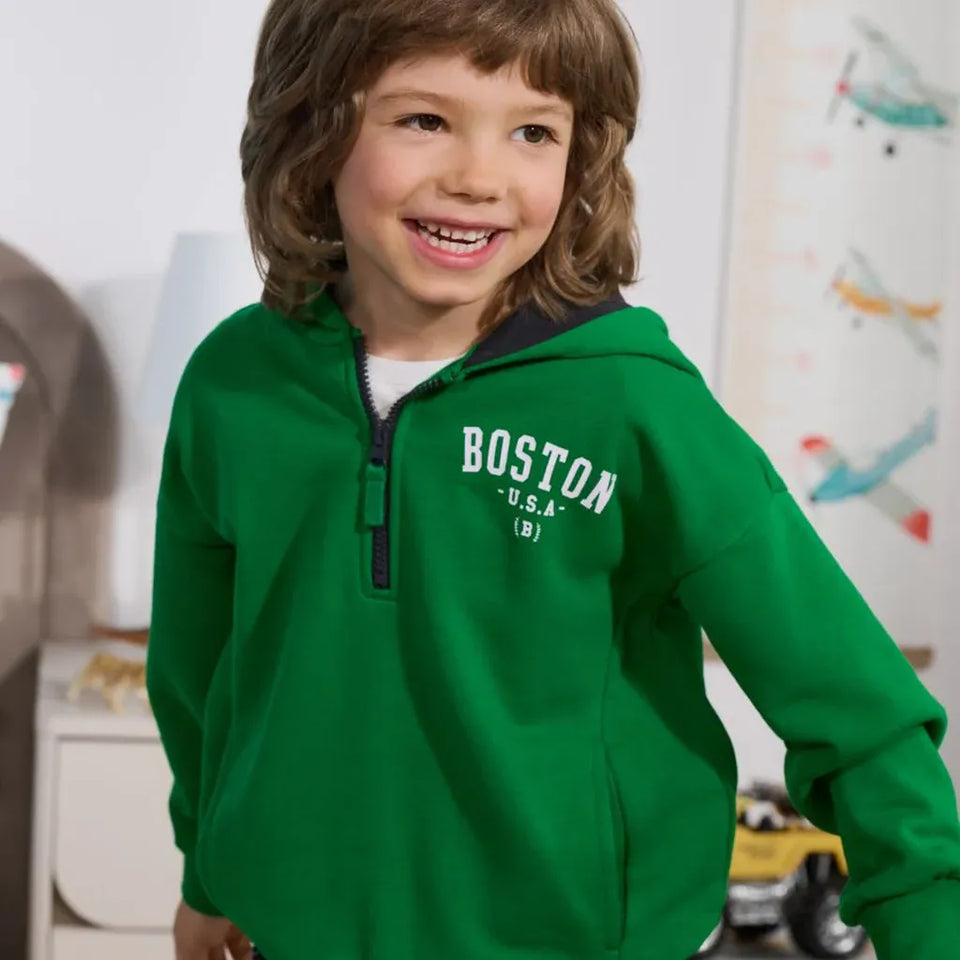 A smiling young boy with brown hair wearing a bright green quarter-zip hoodie with 'BOSTON U.S.