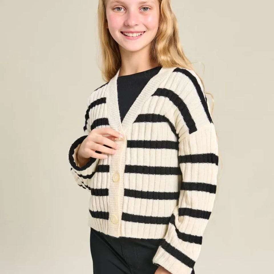 A young girl with blonde hair smiling while wearing a cream and black horizontally striped ribbed knit cardigan over a black top, set against a plain beige studio background.