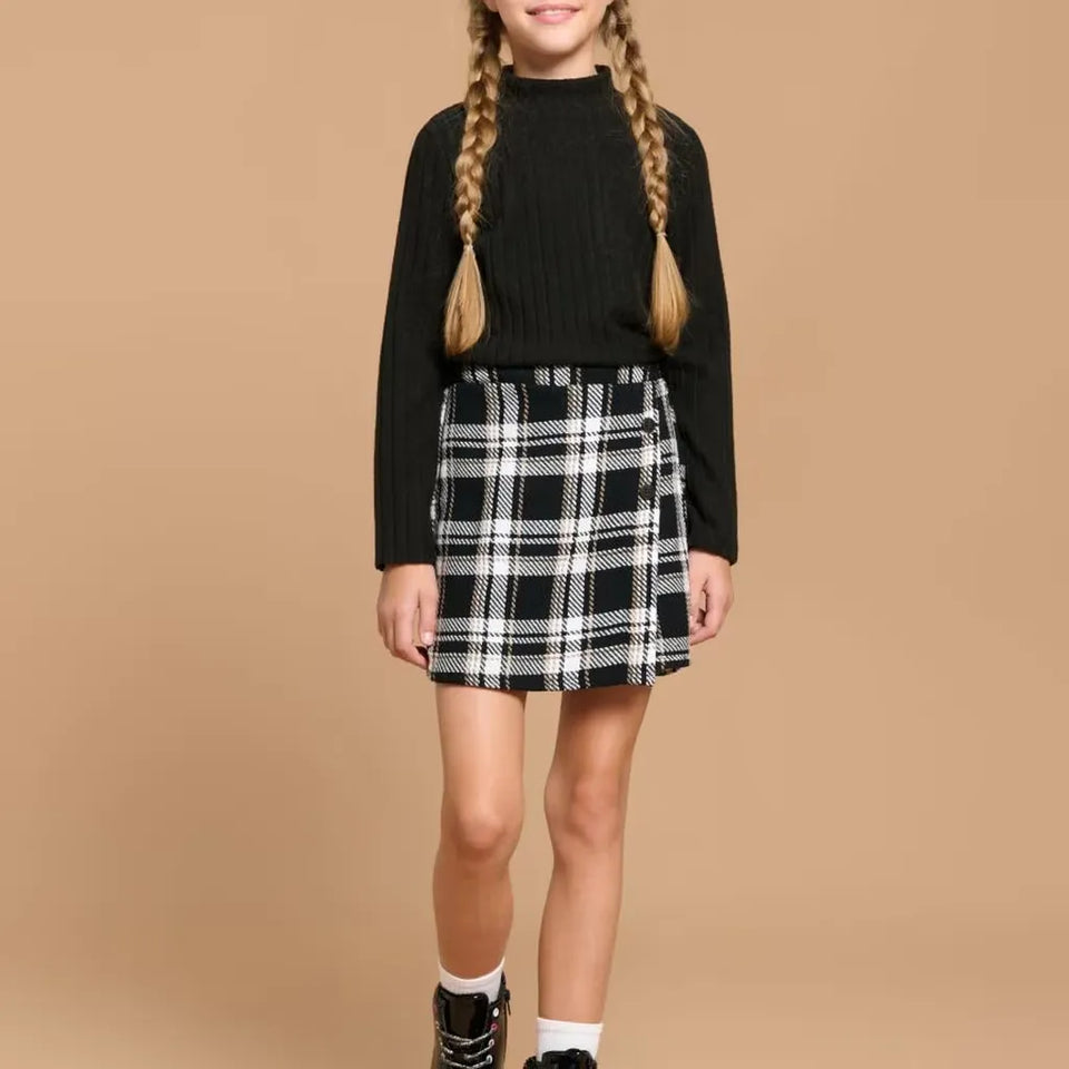 A young girl with long blonde braids is wearing a black ribbed mock-neck long-sleeve sweater and a black and white plaid mini skirt, standing against a solid beige studio background.