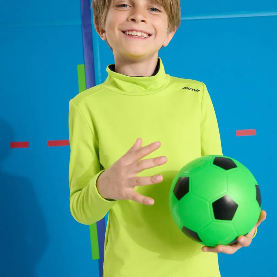 A smiling boy wearing a bright neon yellow-green long-sleeve athletic shirt holds a green soccer ball with black panels against a blue studio background with colorful geometric lines.