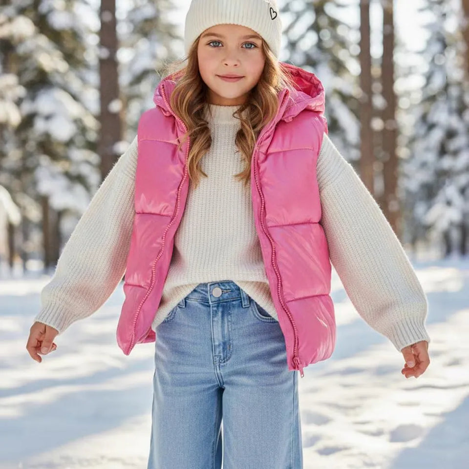A young girl wearing a shiny pink puffer vest over a cream ribbed sweater, blue jeans, and a white knit beanie, standing in a snowy outdoor setting with trees in the background.
