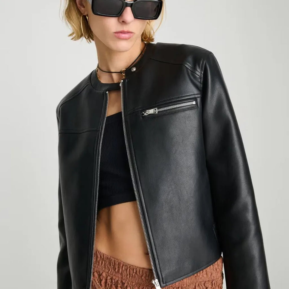 A close-up of a model wearing a black faux-leather moto jacket with a zippered front and a zippered chest pocket, styled with a black crop top and brown textured pants against a light gray background.