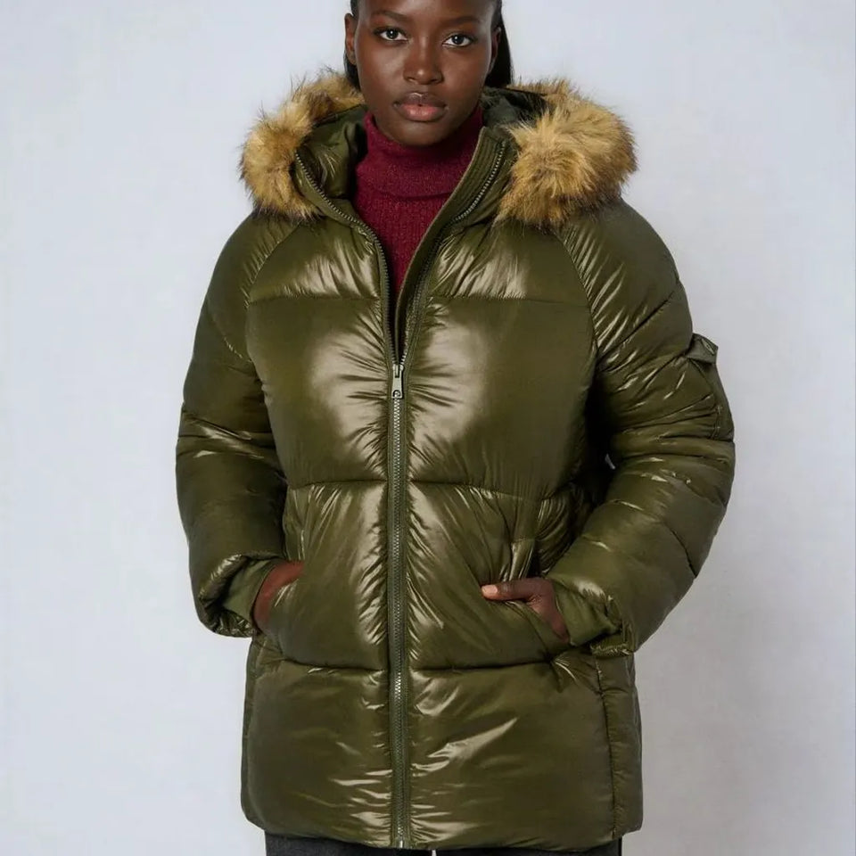 A woman wearing a shiny olive green puffer jacket with a tan faux-fur trimmed hood, layered over a burgundy turtleneck, against a plain light gray studio background.