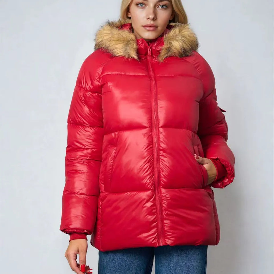 A woman wearing a shiny red puffer jacket with a tan faux-fur trimmed hood, paired with blue jeans, against a light gray studio background.