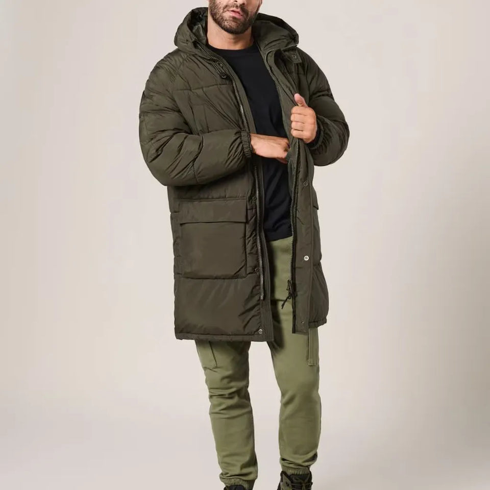 A man wearing a long hooded puffer parka in olive green, styled over a black shirt and olive green pants, standing against a plain light gray studio background.