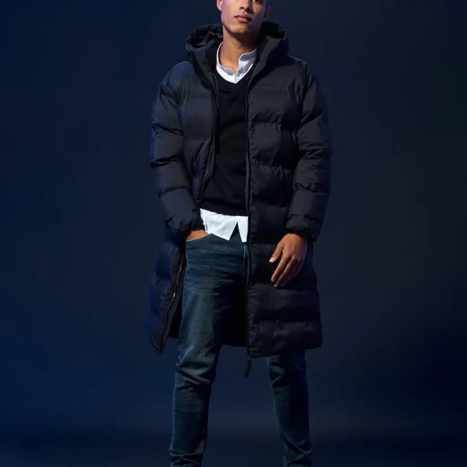 A man wearing a long black hooded puffer jacket over a black sweater and white shirt, paired with dark blue jeans, standing against a dark blue studio background.