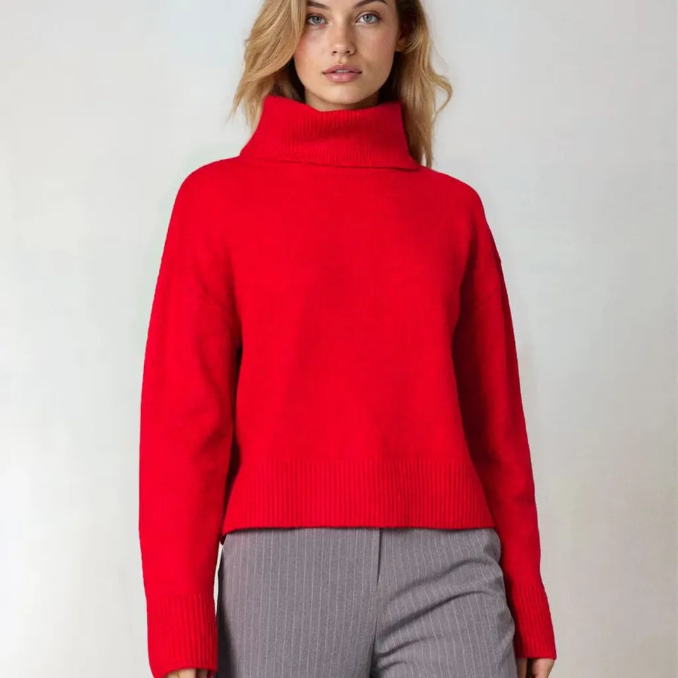 A woman wearing a bright red cropped turtleneck sweater and gray pinstriped trousers, standing against a plain light gray studio background.